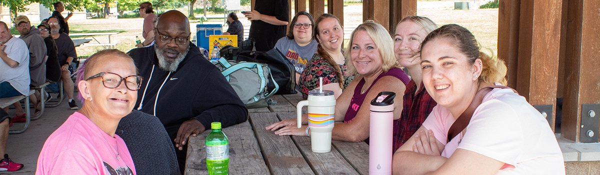 A group from Optimae's Community Integration program sits around a picnic table at a park shelter.